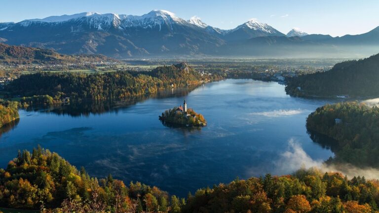 Pohled z lesnaté vyhlídky na Bledské jezero s ostrovem a kostelem, hradem na útesu a Julskými Alpami v pozadí