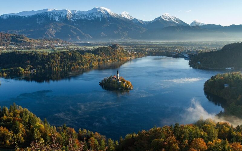 Pohled z lesnaté vyhlídky na Bledské jezero s ostrovem a kostelem, hradem na útesu a Julskými Alpami v pozadí