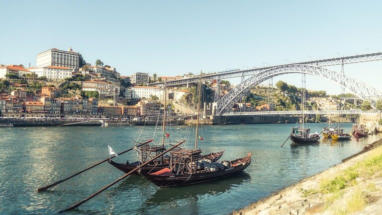 Barevné domy na nábřeží Ribeira v Portu nad řekou Douro, v pozadí železný obloukový most a u břehu dřevěné lodě rabelo