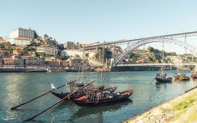 Barevné domy na nábřeží Ribeira v Portu nad řekou Douro, v pozadí železný obloukový most a u břehu dřevěné lodě rabelo