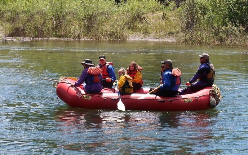Prázdný červený nafukovací raft s pádly, vestami a přilbami na kamenitém břehu horské řeky s peřejemi