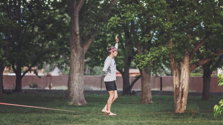 Nízko napnutá slackline mezi dvěma silnými stromy v parku s ochranou kůry pod smyčkami a nohy člověka balancujícího na popruhu