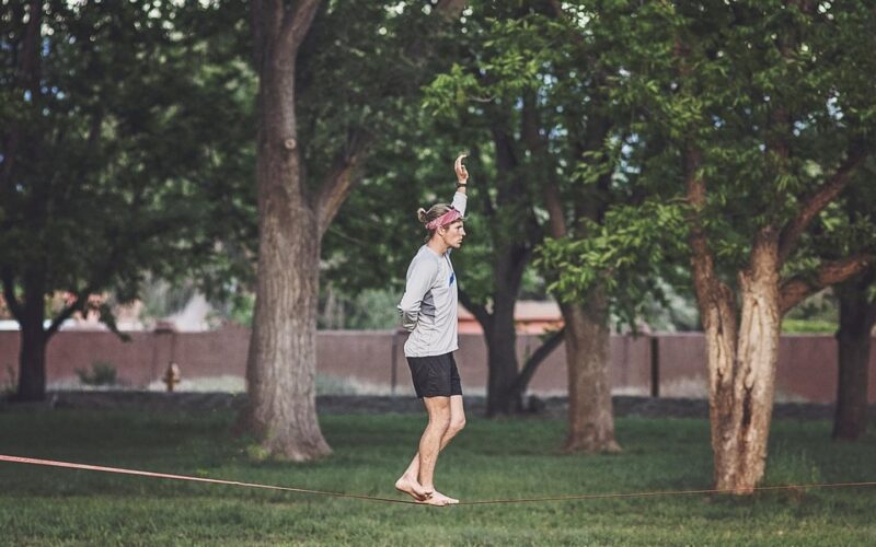 Nízko napnutá slackline mezi dvěma silnými stromy v parku s ochranou kůry pod smyčkami a nohy člověka balancujícího na popruhu